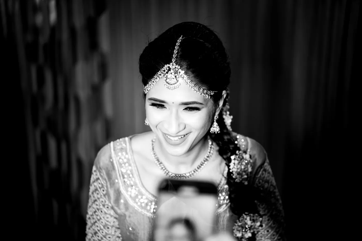 Black and white of Medha laughing softly in a headpiece while a phone is held in front of her