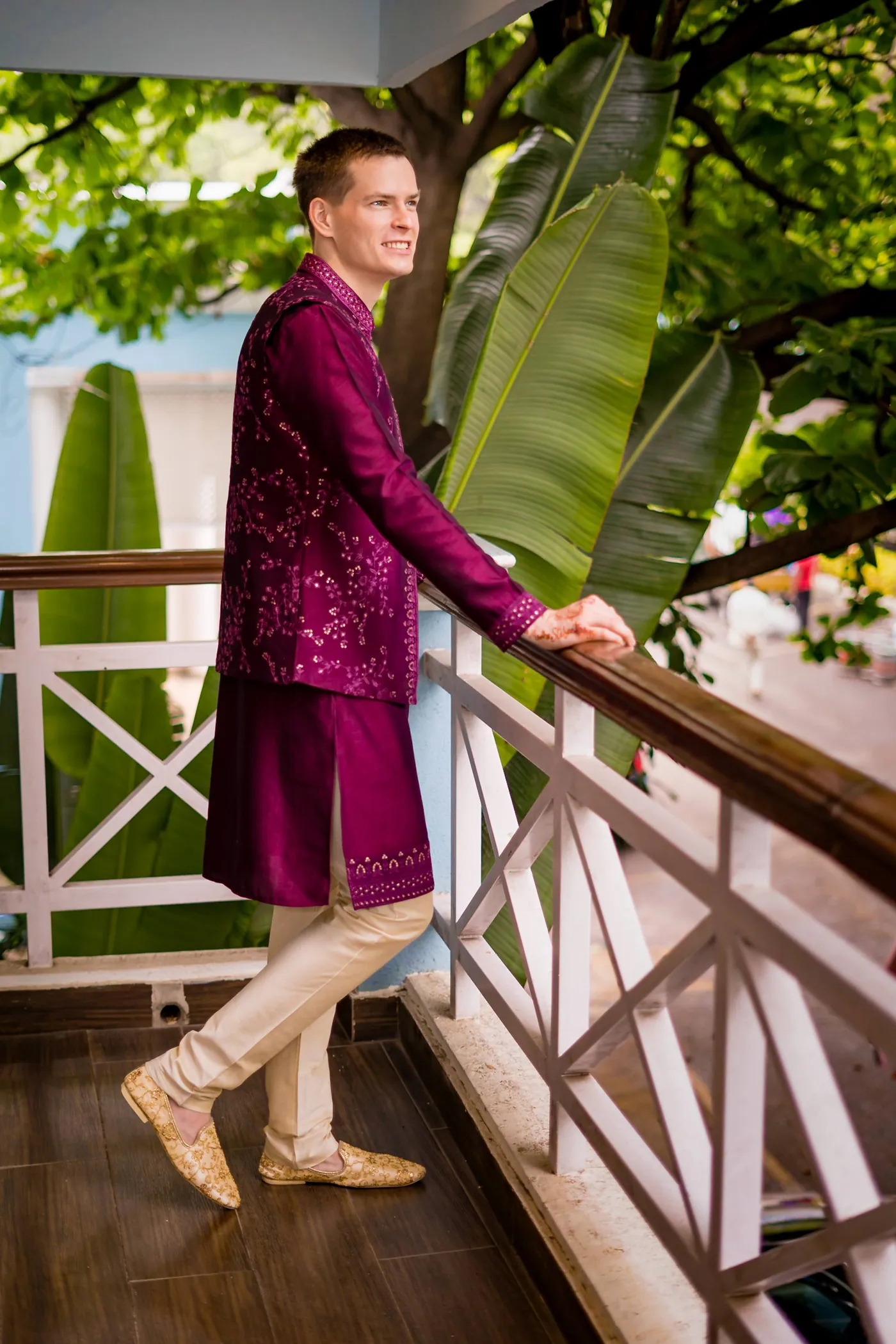 Stefan leans on a white balcony railing in a magenta jacket under large banana leaves
