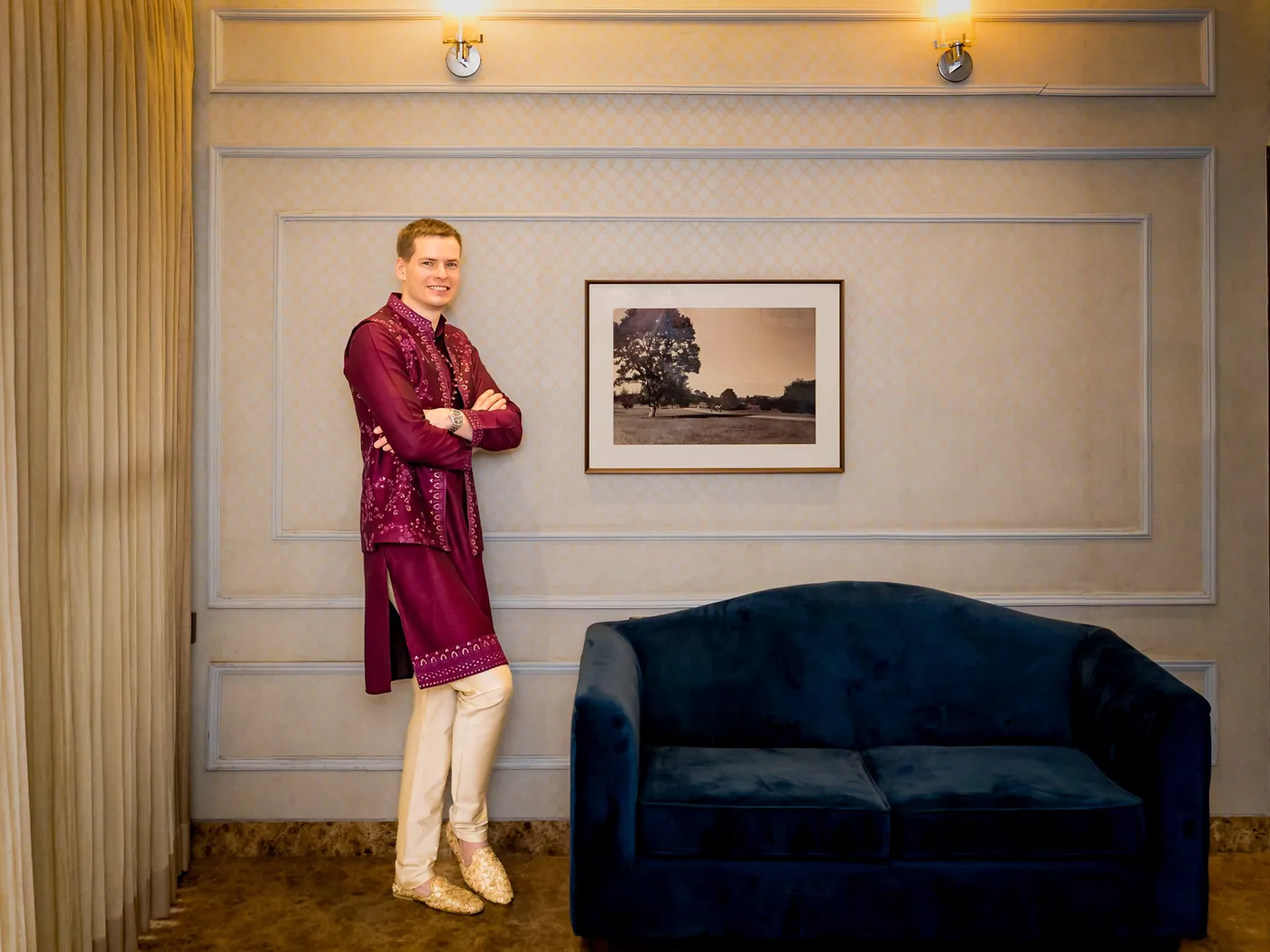 Stefan stands with arms crossed against a cream wall beside a blue sofa and a framed picture