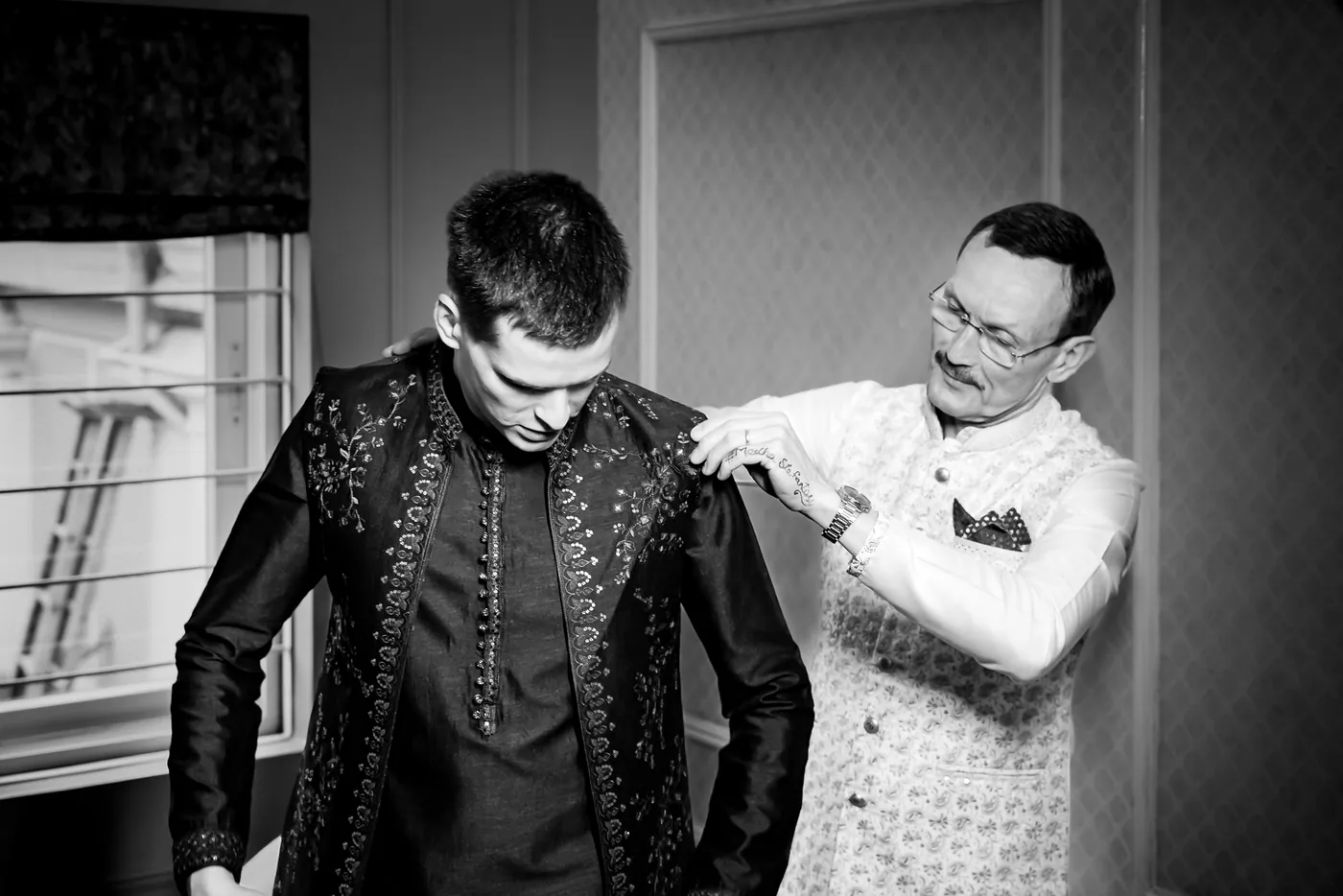 Stefan's father in a cream waistcoat straightens the shoulder of Stefan's dark embroidered jacket by a window
