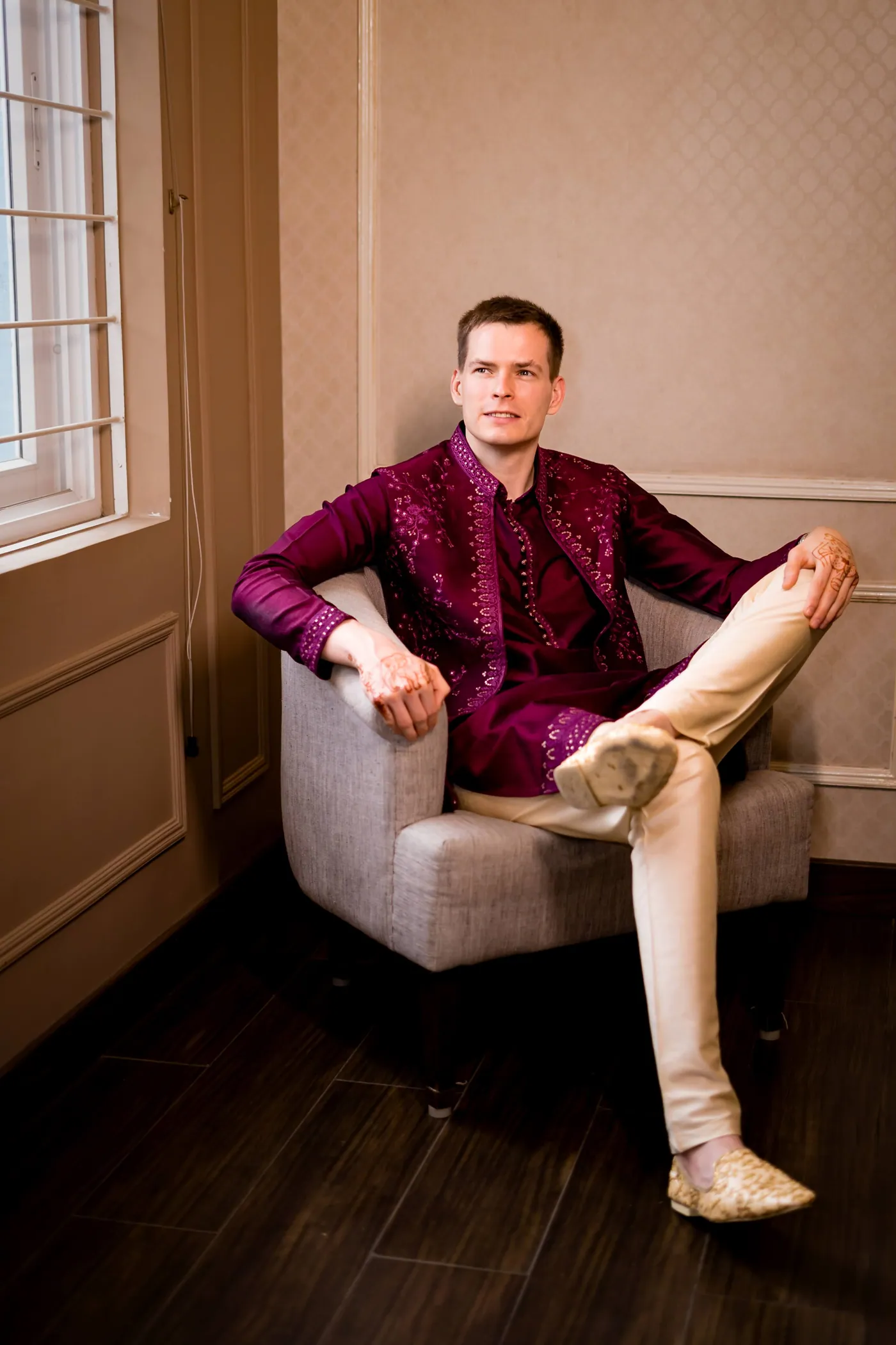 Stefan in a magenta kurta lounges in an armchair by a window with one leg crossed