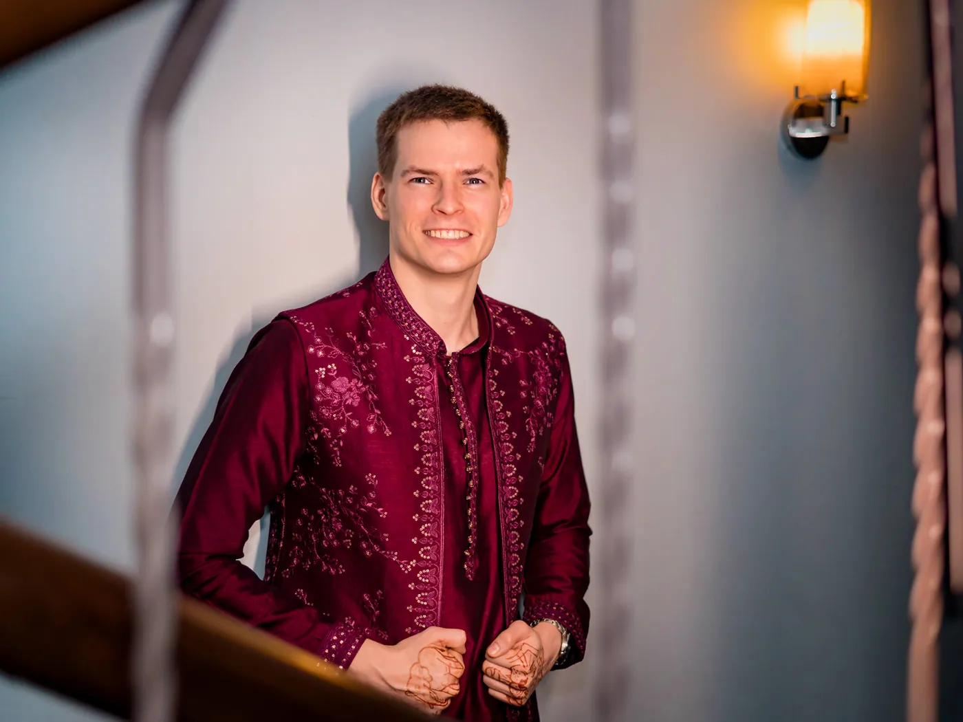 Stefan in a magenta embroidered jacket stands on a staircase landing beside a warm wall lamp