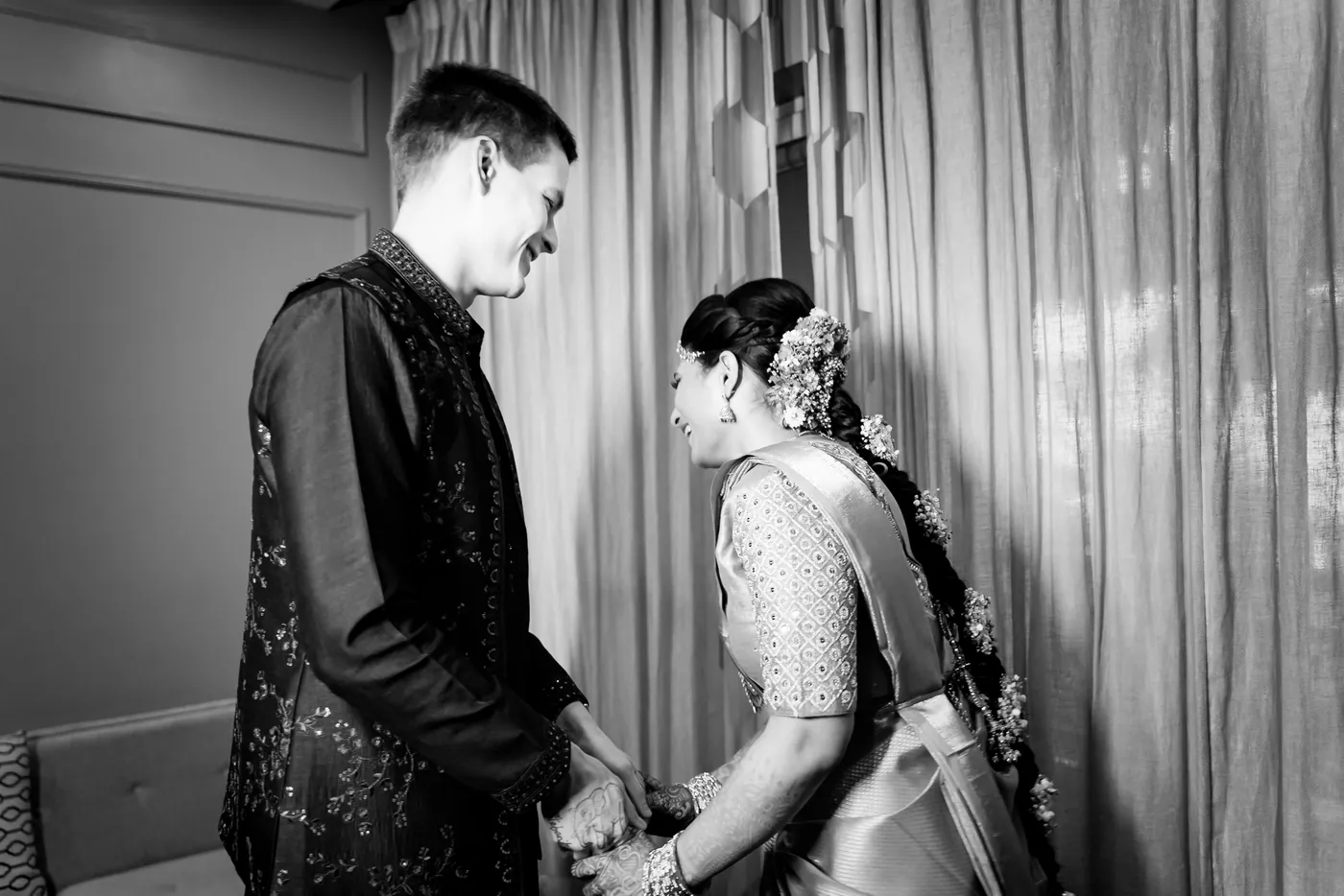 Black and white of Stefan and Medha holding hands and smiling at each other by a curtained window