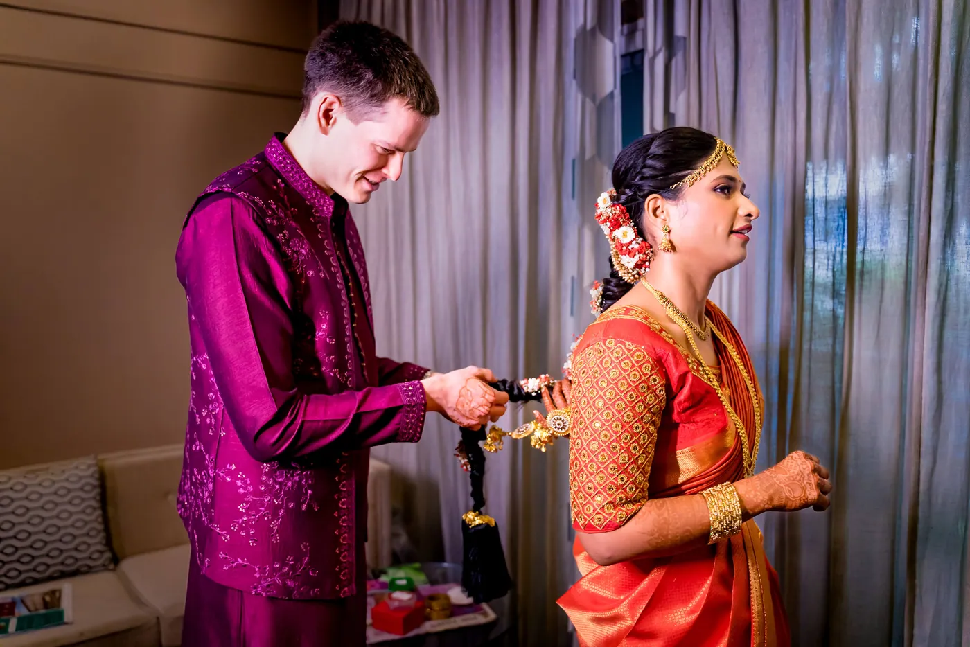 Stefan holds a small gold piece near Medha as she smiles sideways at him by a curtain
