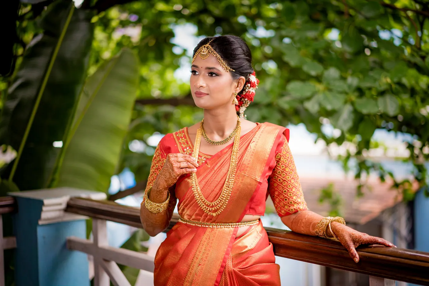 Medha in an orange silk sari leans on a balcony railing with green banana leaves behind