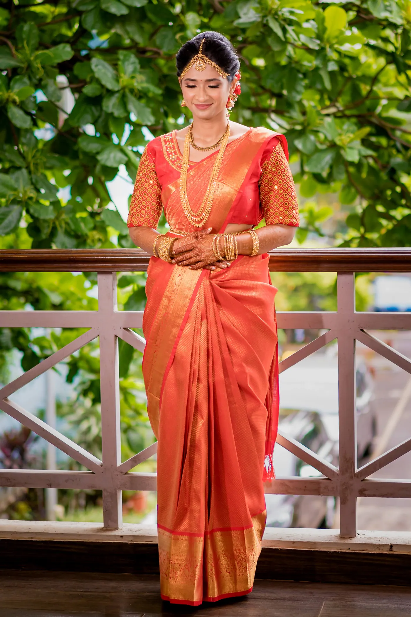 Medha stands with folded hands on a wooden balcony in a full-length orange silk sari