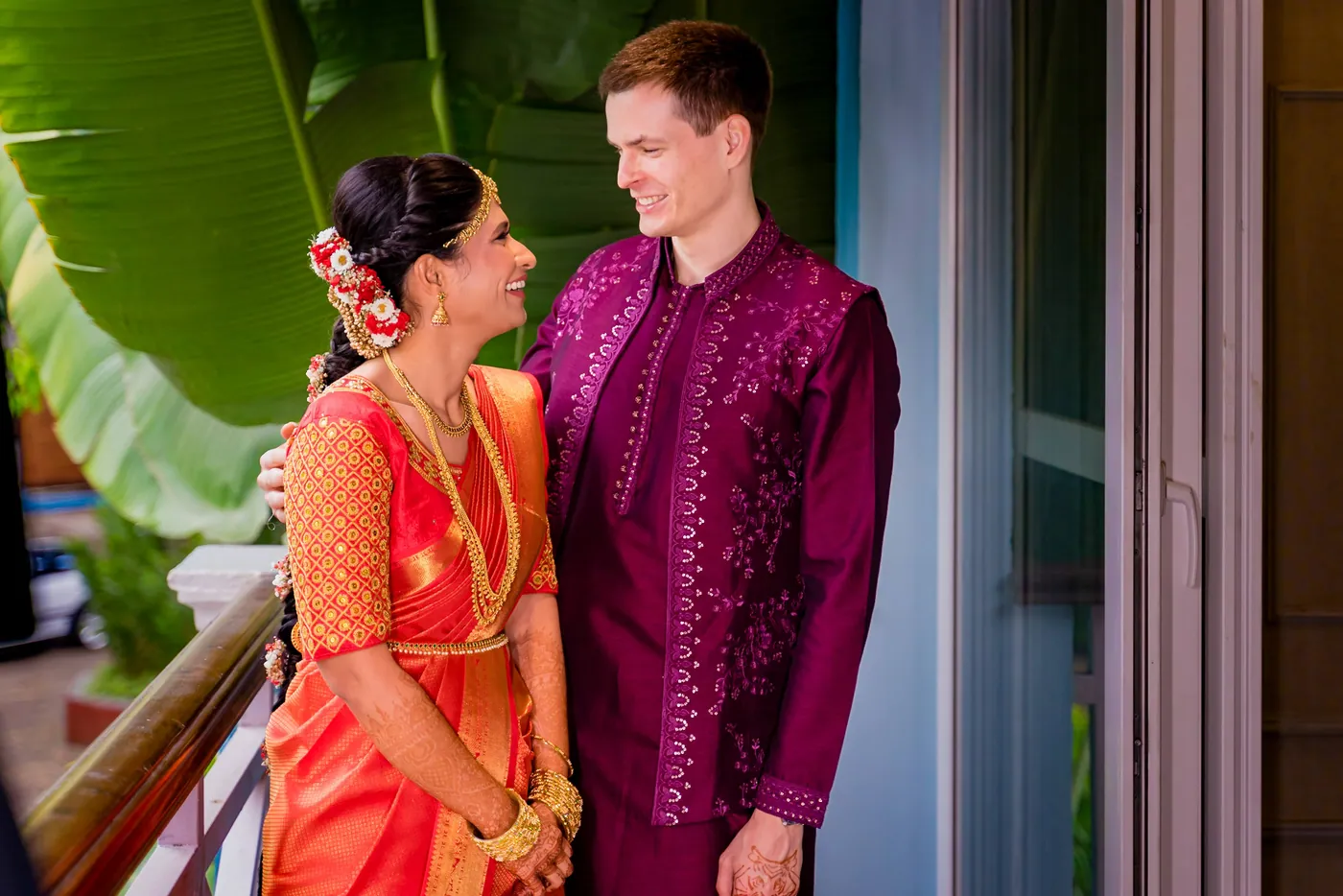 Medha and Stefan smile at each other on the balcony, she in orange and he in magenta