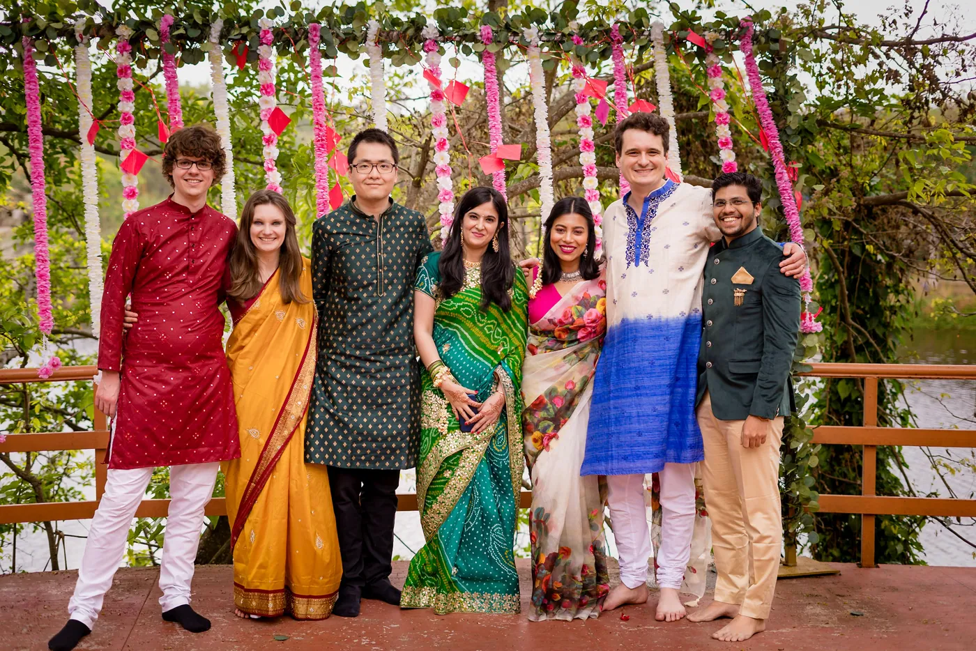Seven friends in colorful traditional clothes pose in front of hanging flowers
