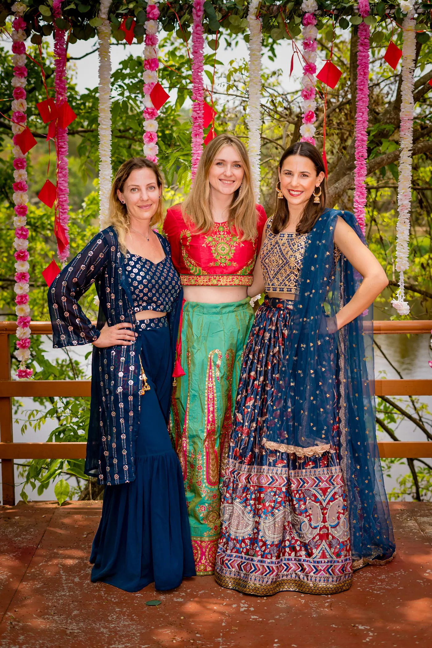 Three women in blue and green traditional clothes pose in front of a flower backdrop
