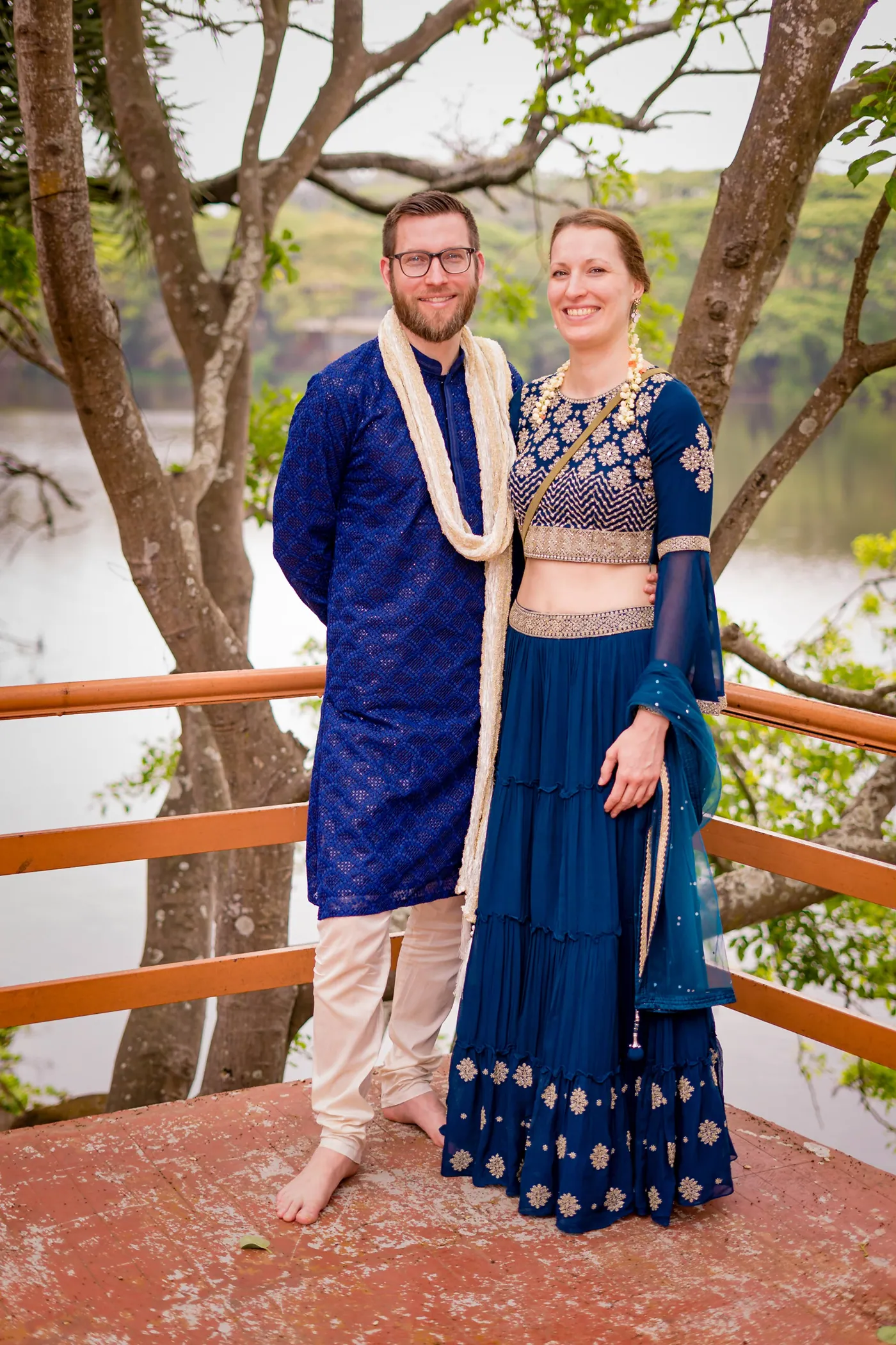 A man in a blue kurta and a woman in a blue skirt stand on a deck above a lake