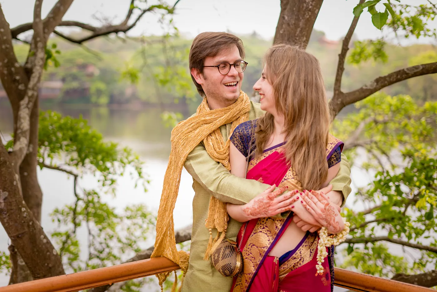 A man in a golden shawl and a woman in a red sari hug beside a lake