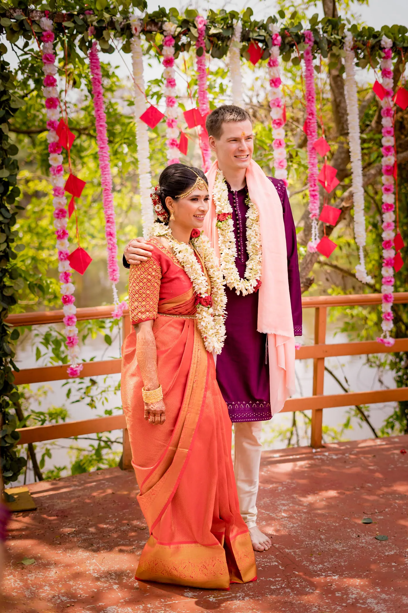 Medha and Stefan pose in garlands in front of hanging pink and white flowers on the deck