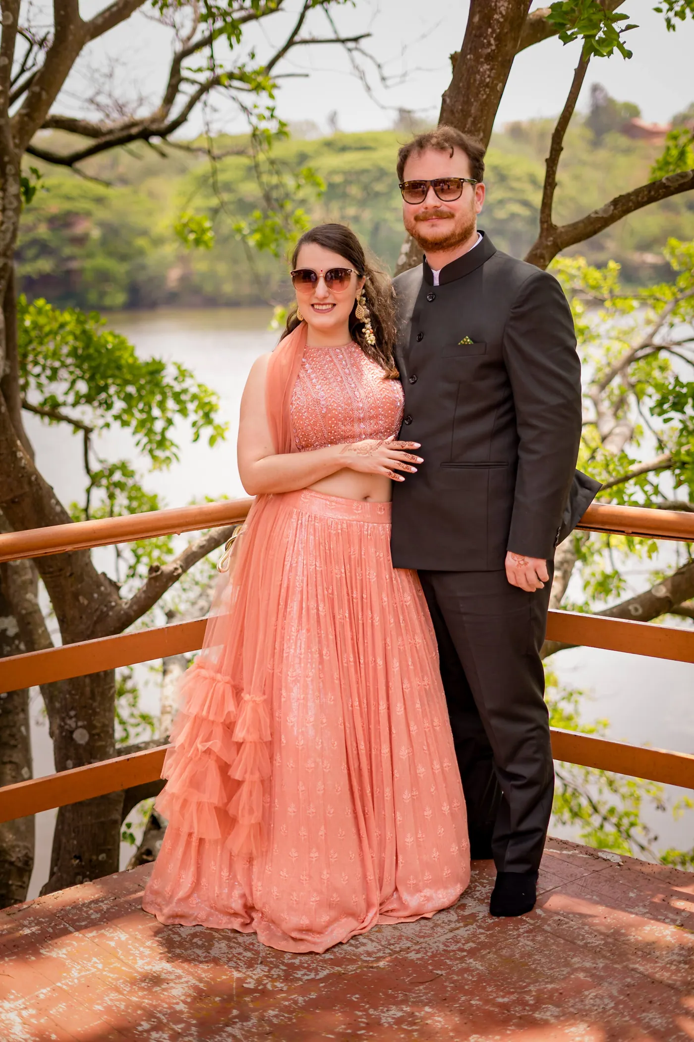 A woman in a peach gown and a man in a black suit pose at a lake railing