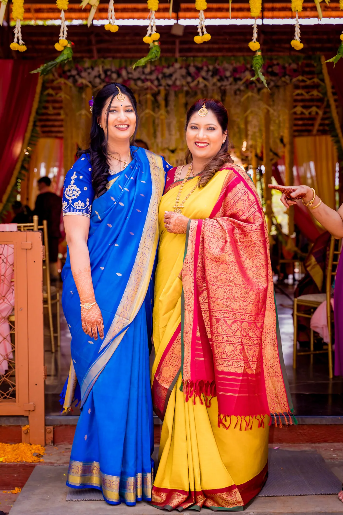 Two women in blue and yellow saris pose together in a warm wooden hall