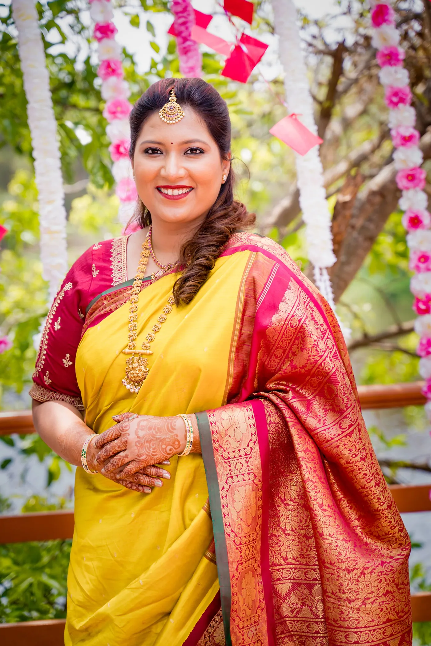 A woman in a yellow and red sari smiles in front of hanging flowers