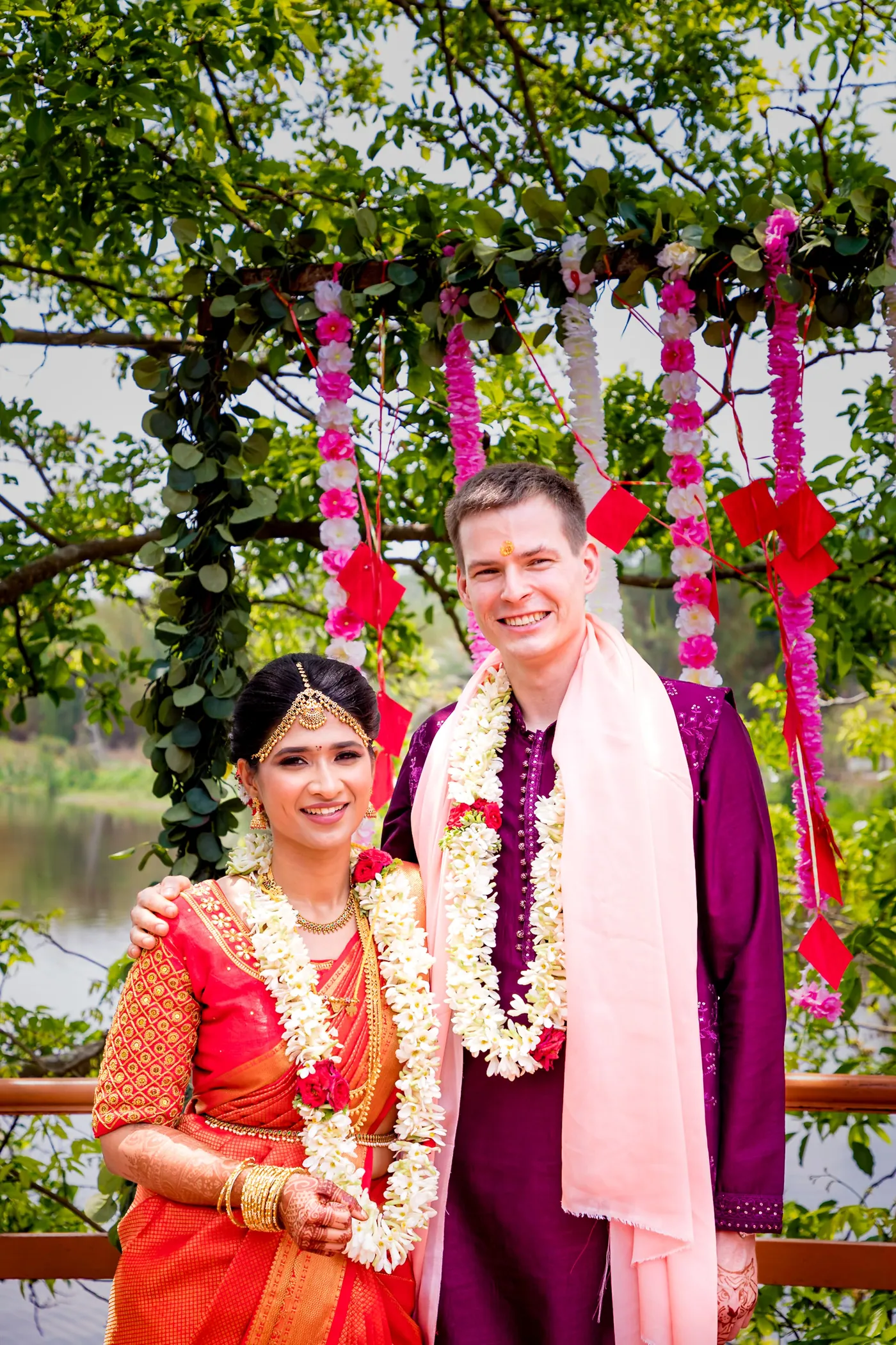 Medha and Stefan wearing flower garlands pose smiling at a wooden rail with hanging pink flower strings
