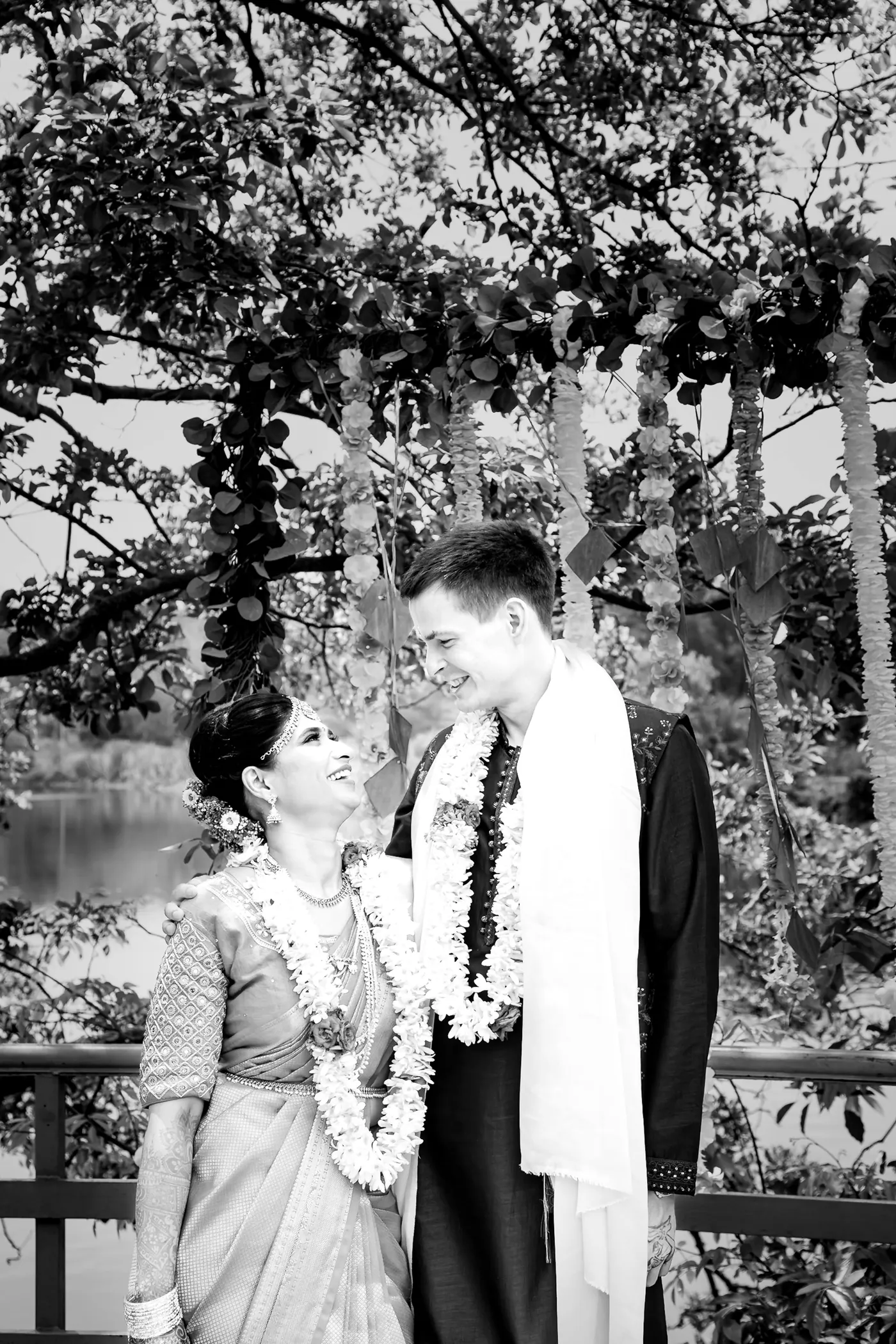 Black and white of Medha in a garland smiling up at Stefan beside a lakeside railing