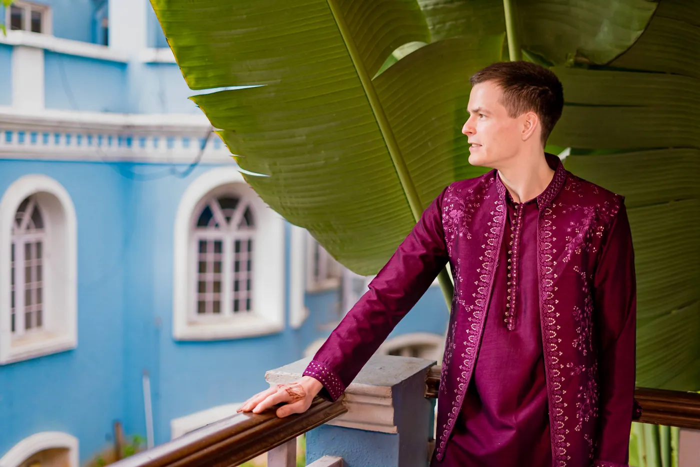 Stefan in a magenta kurta rests a hand on a balcony rail near banana leaves and a blue building