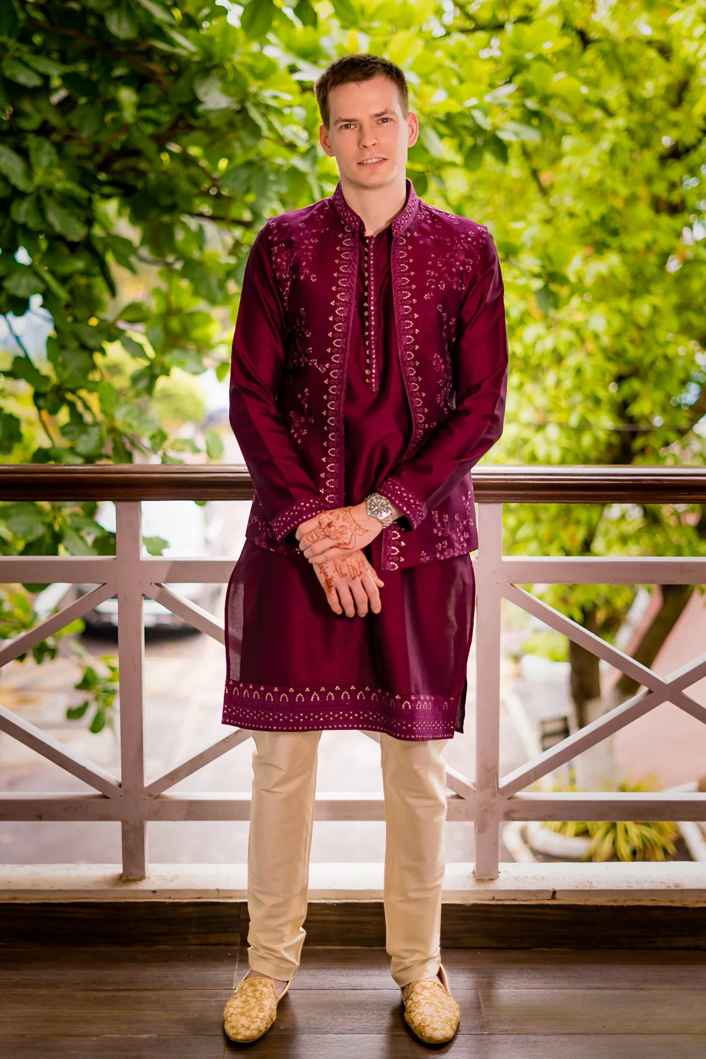 Stefan stands on a wooden balcony in a magenta kurta and cream trousers with green trees behind him