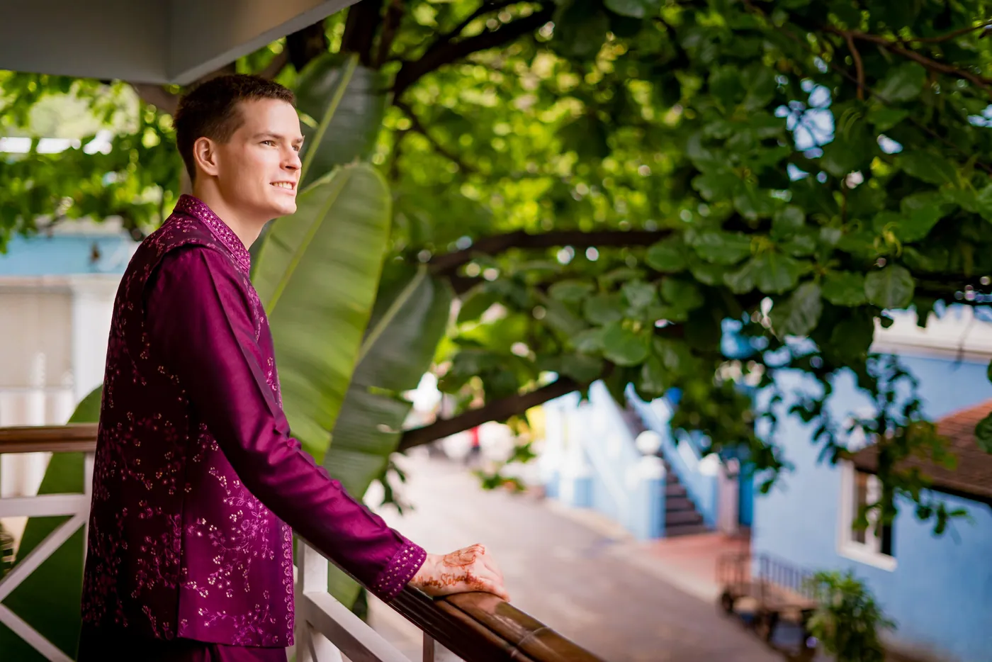 Stefan leans on a balcony rail in a magenta kurta looking away past banana leaves