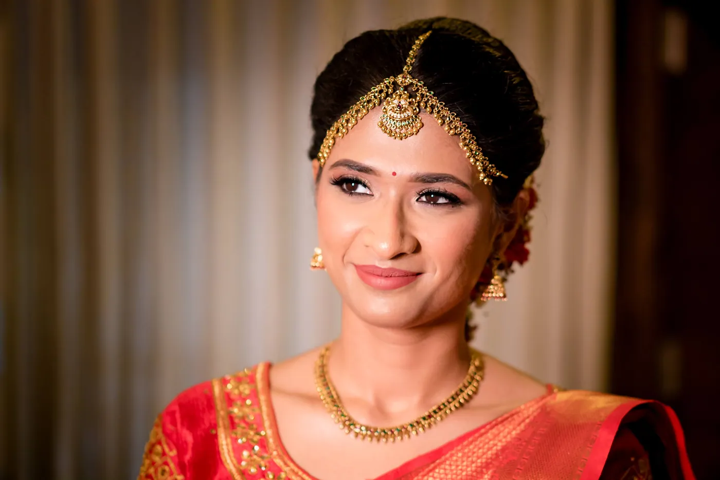 Medha smiles softly in an orange blouse, gold headpiece and necklace, framed against curtains