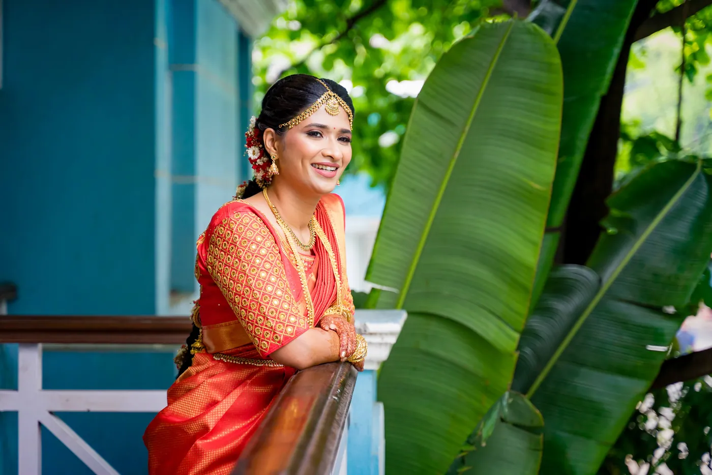 Medha in an orange sari smiles over a wooden rail against large banana leaves and a blue wall