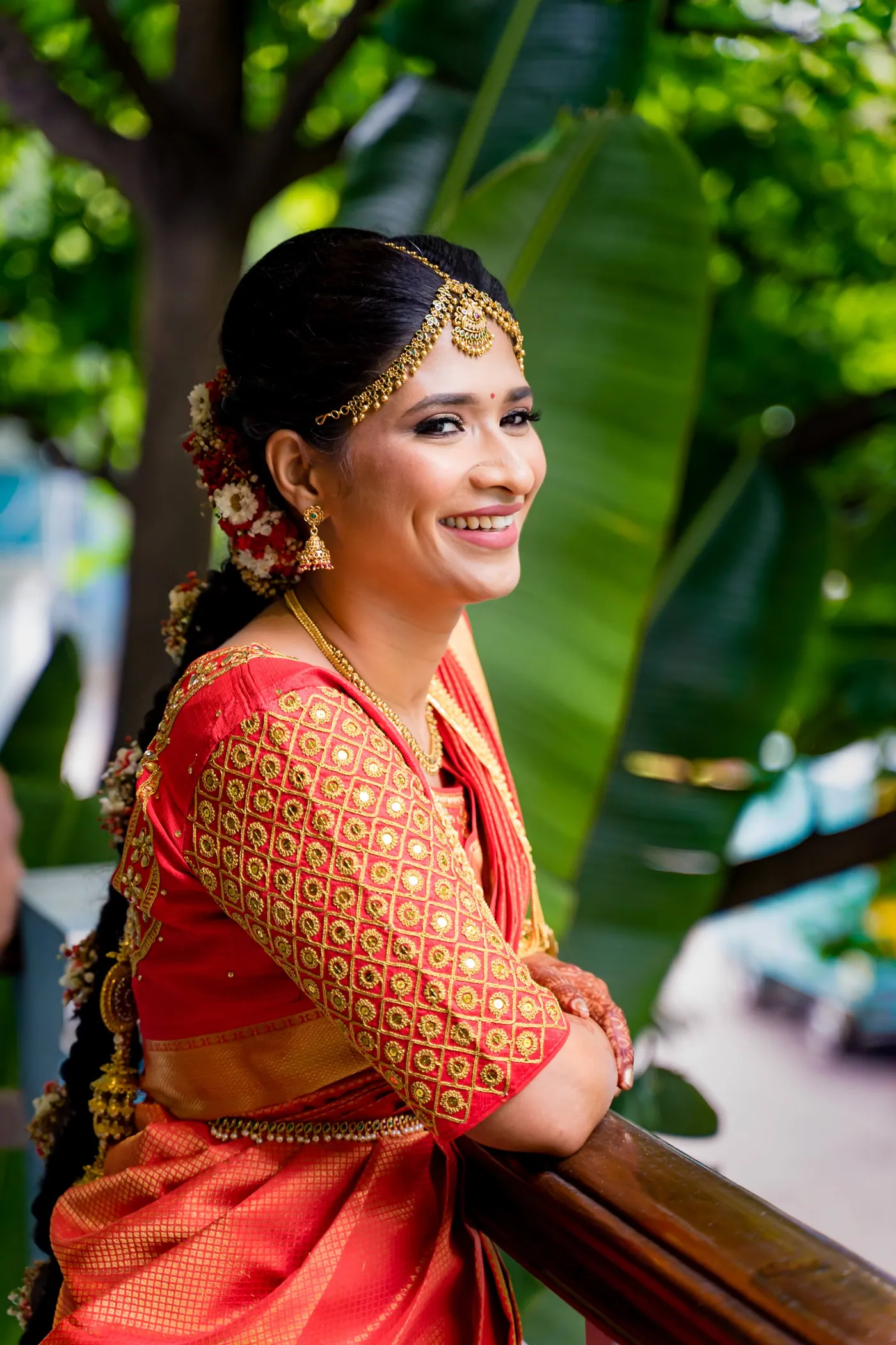 Medha leans on a balcony rail in an orange silk sari with a gold headpiece, smiling at the camera