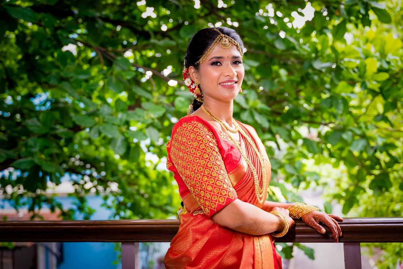 Medha in an orange sari smiles at the camera under green tree branches on a balcony