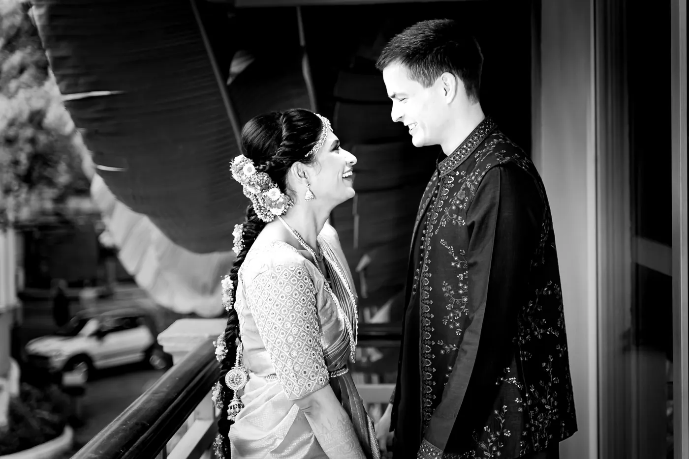 Black and white of Medha and Stefan smiling at each other on a balcony with leaf shadow overhead