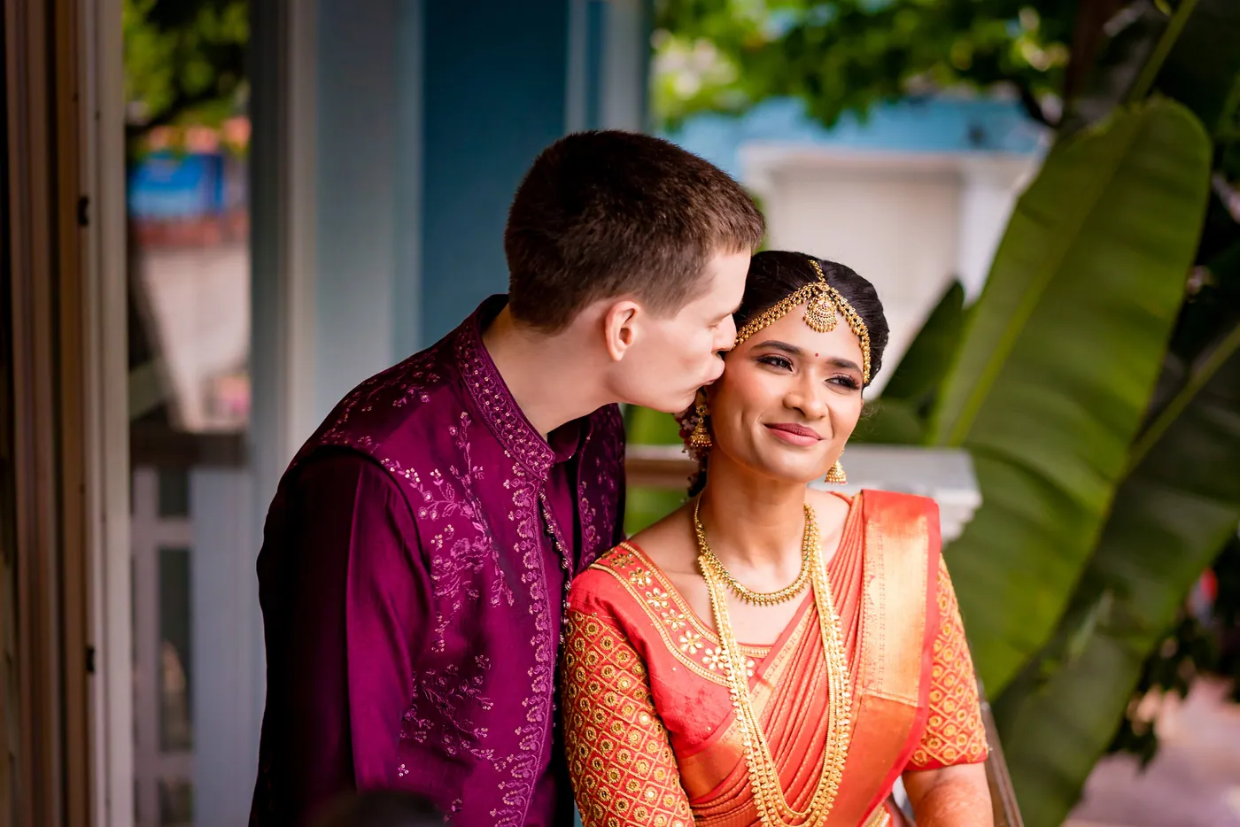 Stefan kisses Medha's cheek as she smiles toward the camera on a balcony