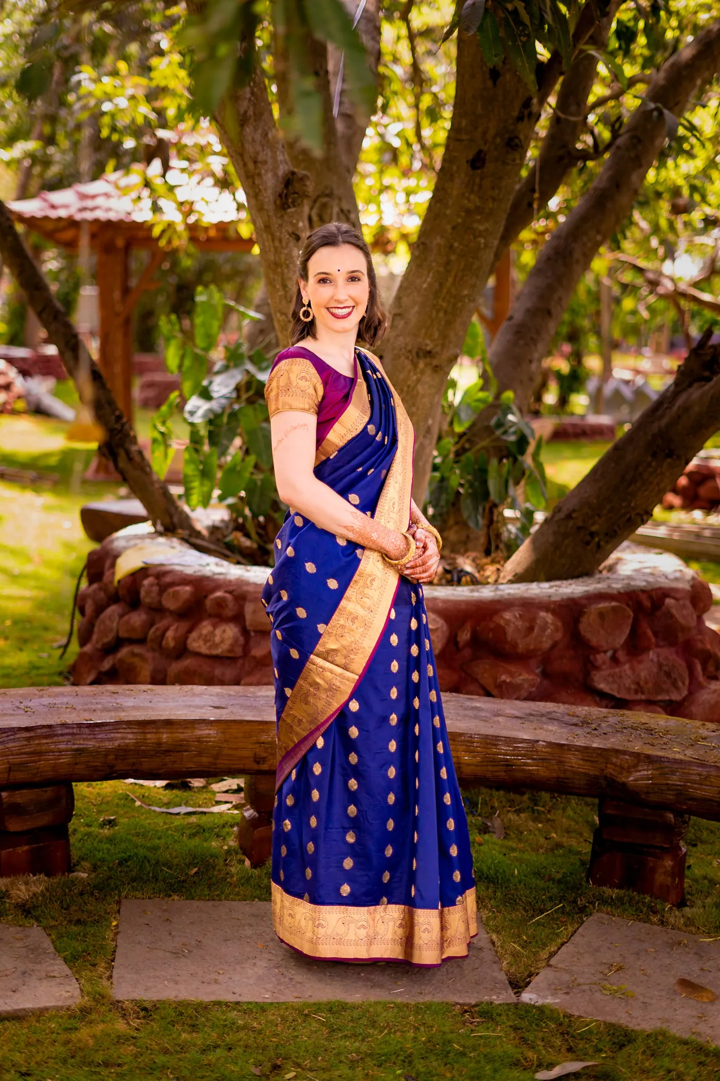 Stefan's sister stands in a royal blue sari with a gold border by a carved log bench in a garden