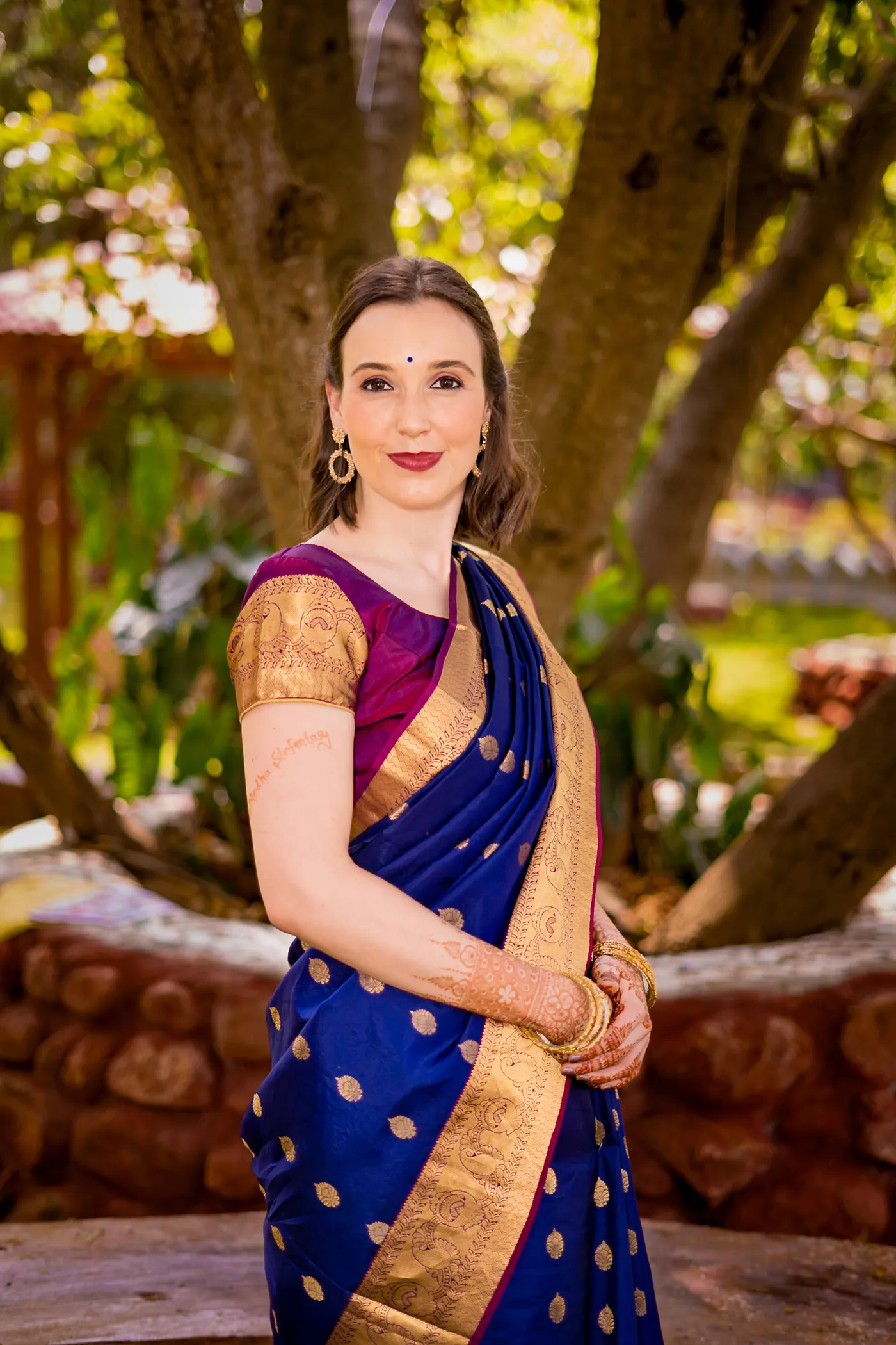 Stefan's sister in a blue and gold sari smiles at the camera beneath a large tree trunk