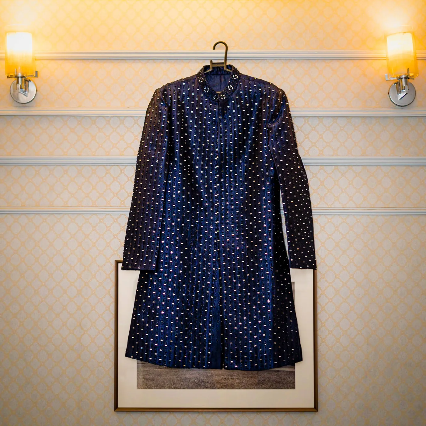 A navy sherwani with a scattered dot pattern hangs on a cream wall between two wall lamps