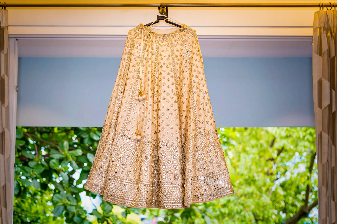 A cream embroidered lehenga hangs in a doorway with green foliage showing behind it