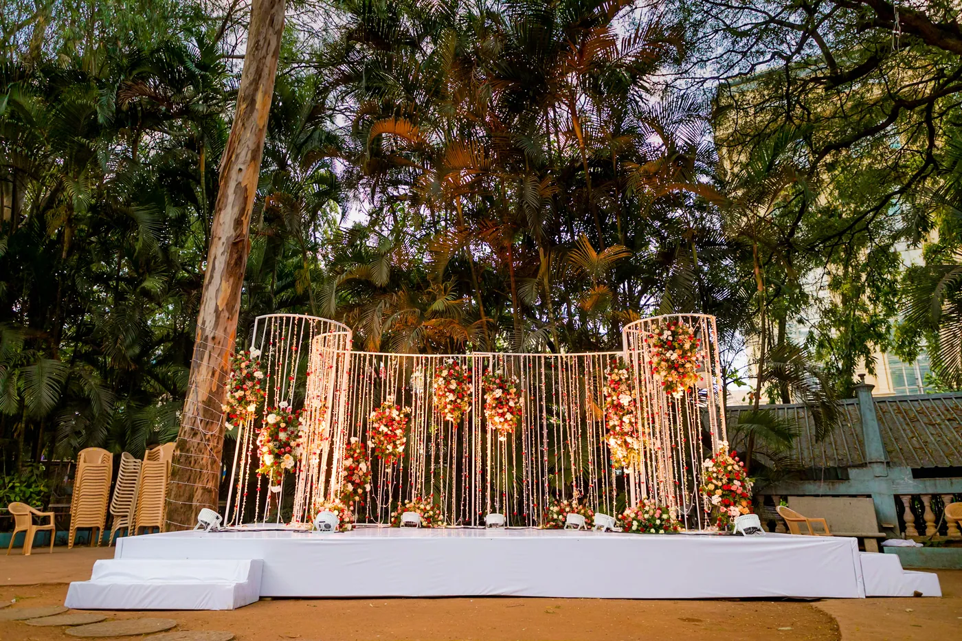 A white stage backed by gold hanging strands is set in a garden clearing between tall trees