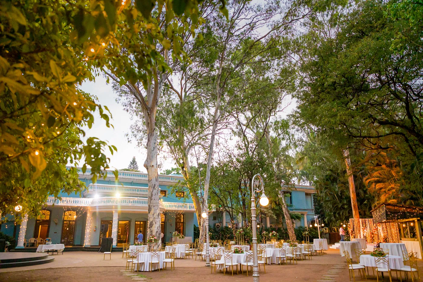 Round tables and soft lights are set under tall trees in front of the blue and white club building