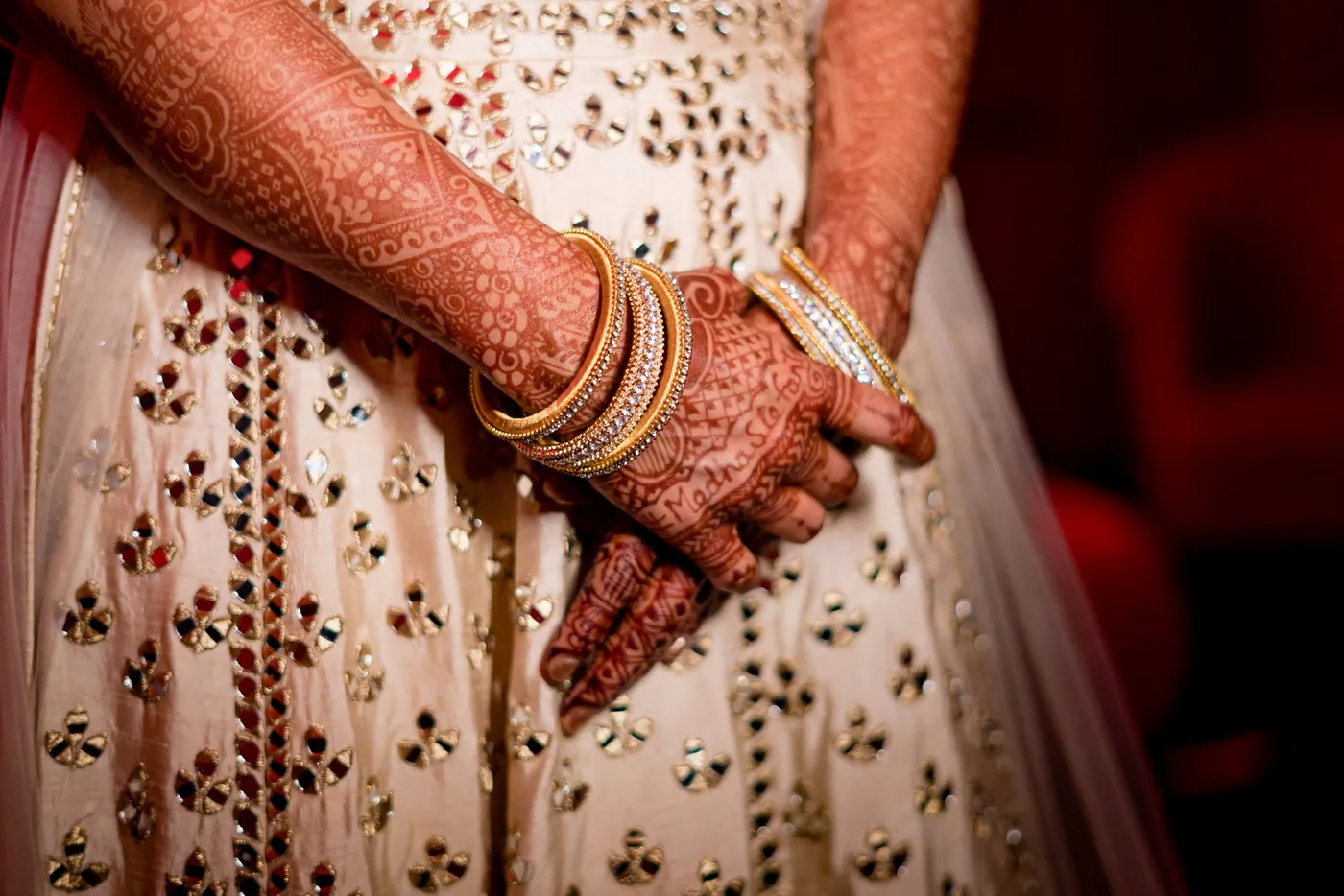 Henna covered hands and gold bangles rest on a cream lehenga embroidered with small mirrors