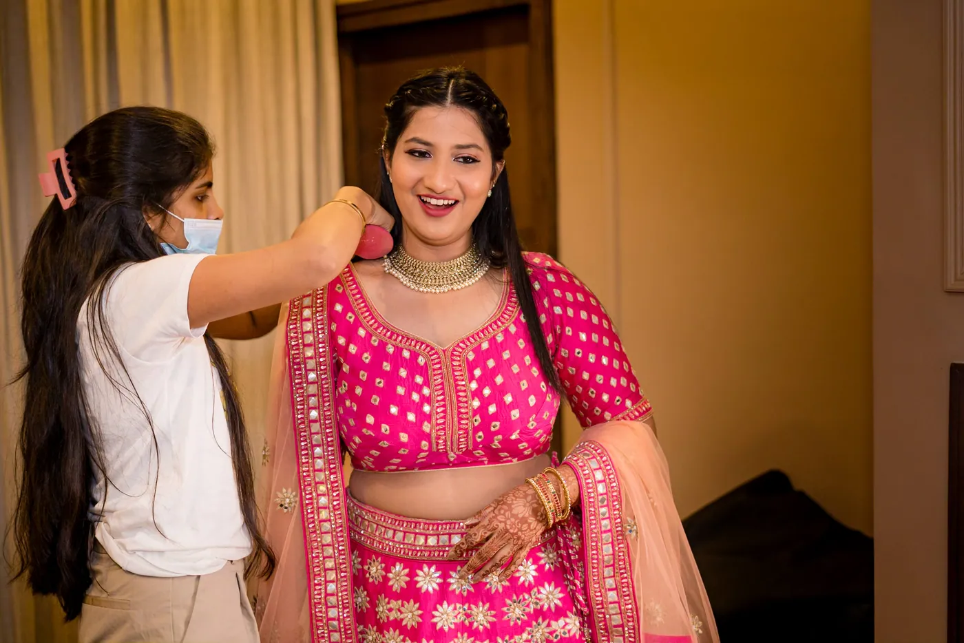 Medha laughs in a pink lehenga while a woman in a white shirt adjusts her dupatta