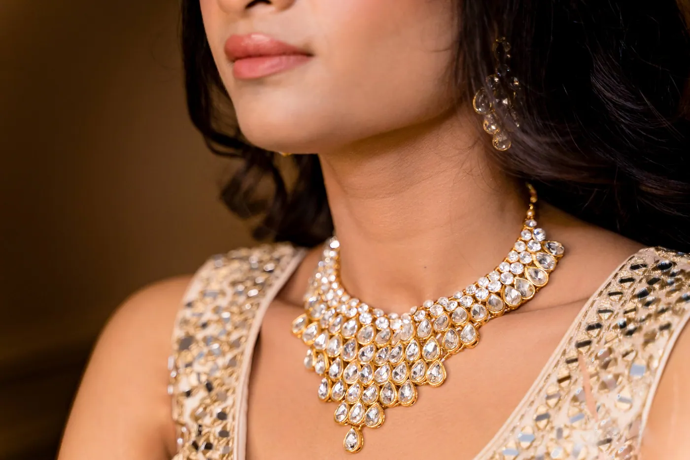 A close crop of a gold and crystal necklace on Medha's collarbone with a soft smile above it