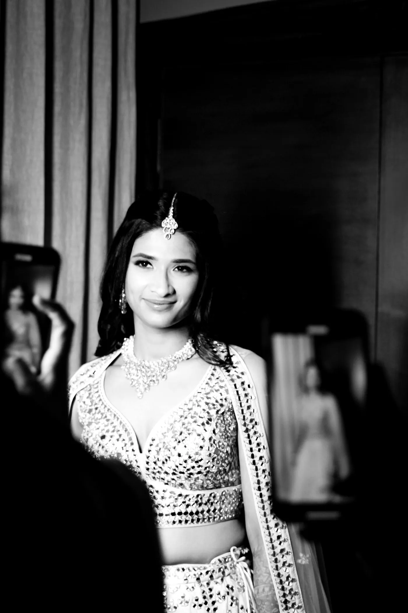 In black and white, Medha in a mirrored blouse stands by a dark curtain with a phone raised behind her