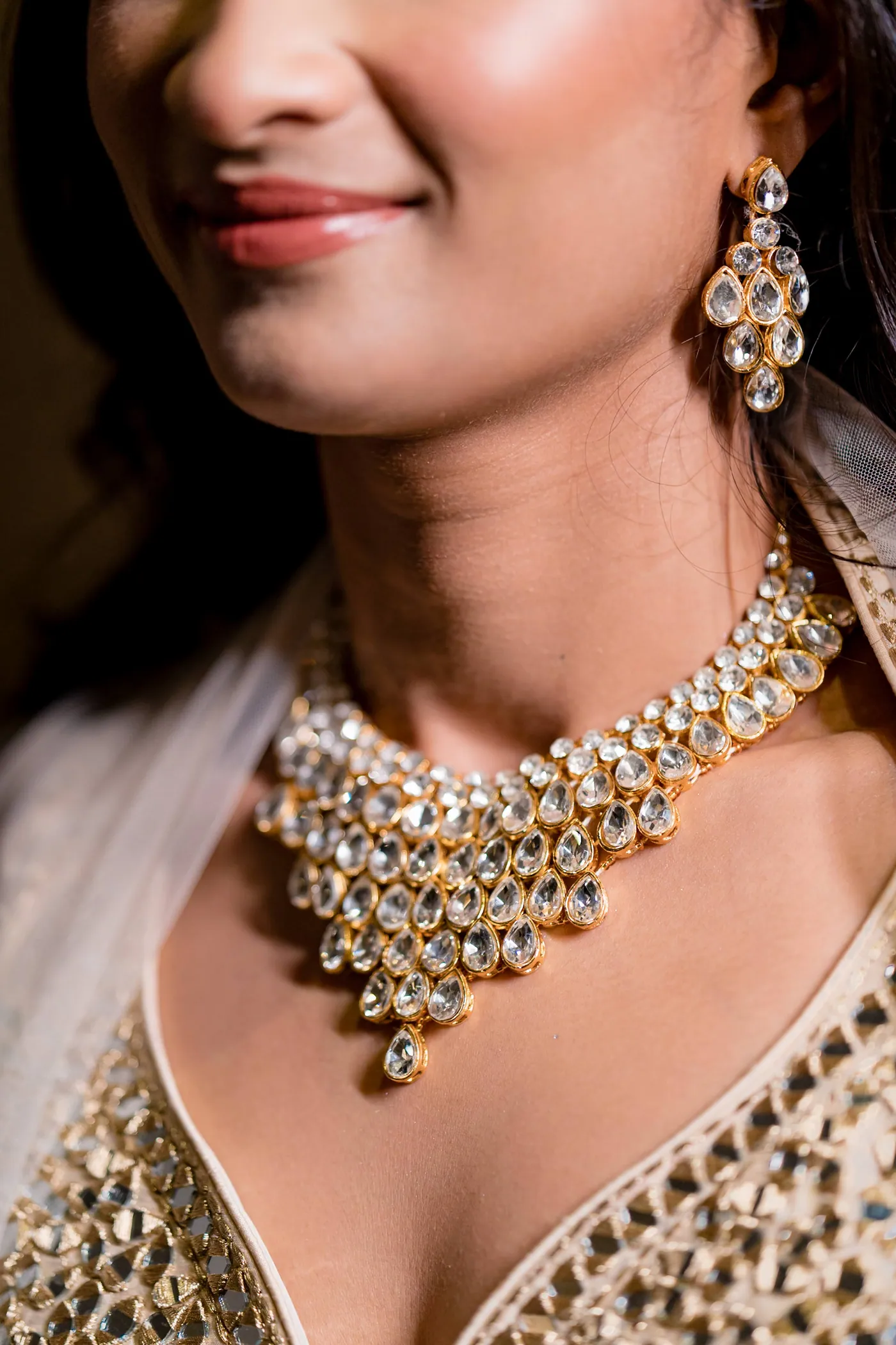A tight crop of Medha's smile, a drop earring and a layered gold necklace