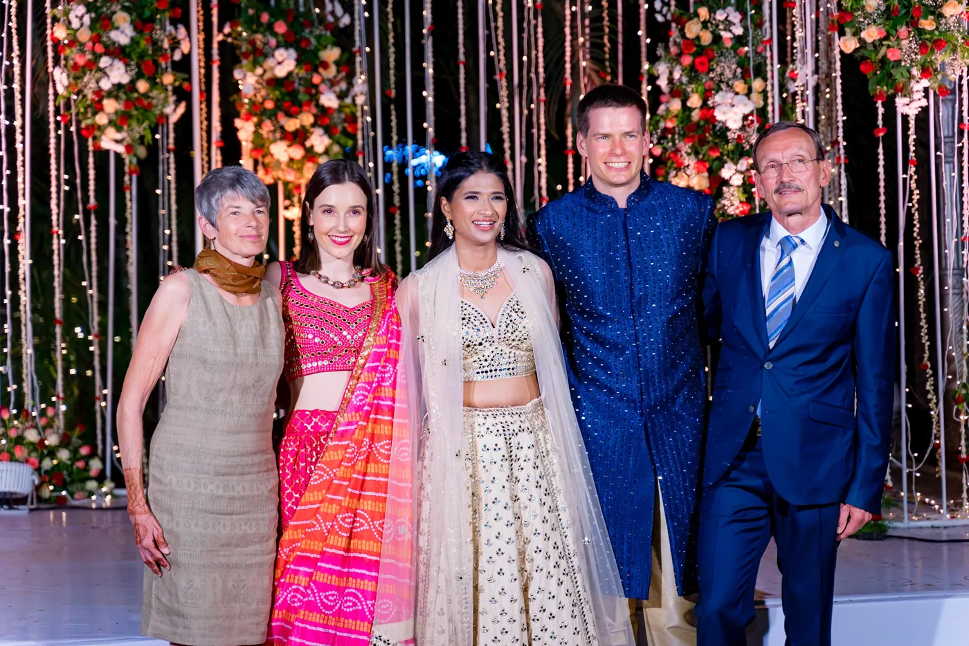 Medha and Stefan stand in a row with family in front of the hanging lights backdrop