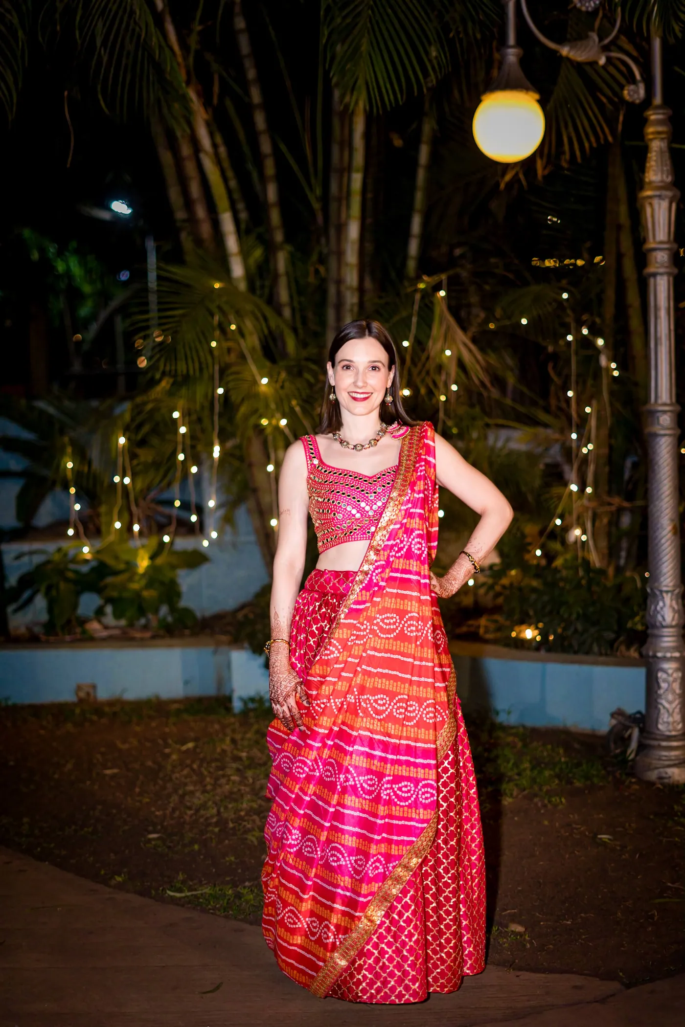 Stefan's sister stands with her hands on her hips in a pink and orange lehenga