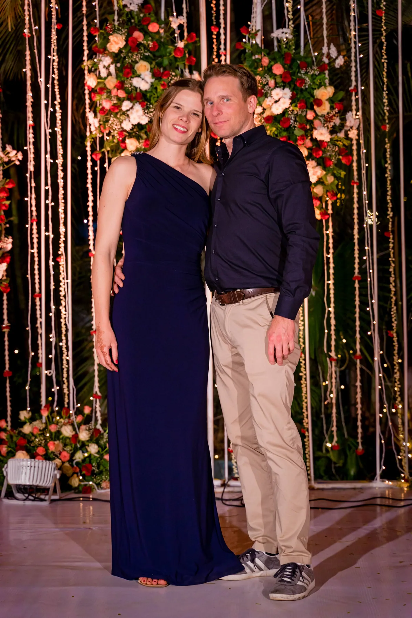 A woman in a navy dress and a man in a dark shirt pose close together before flowers and lights