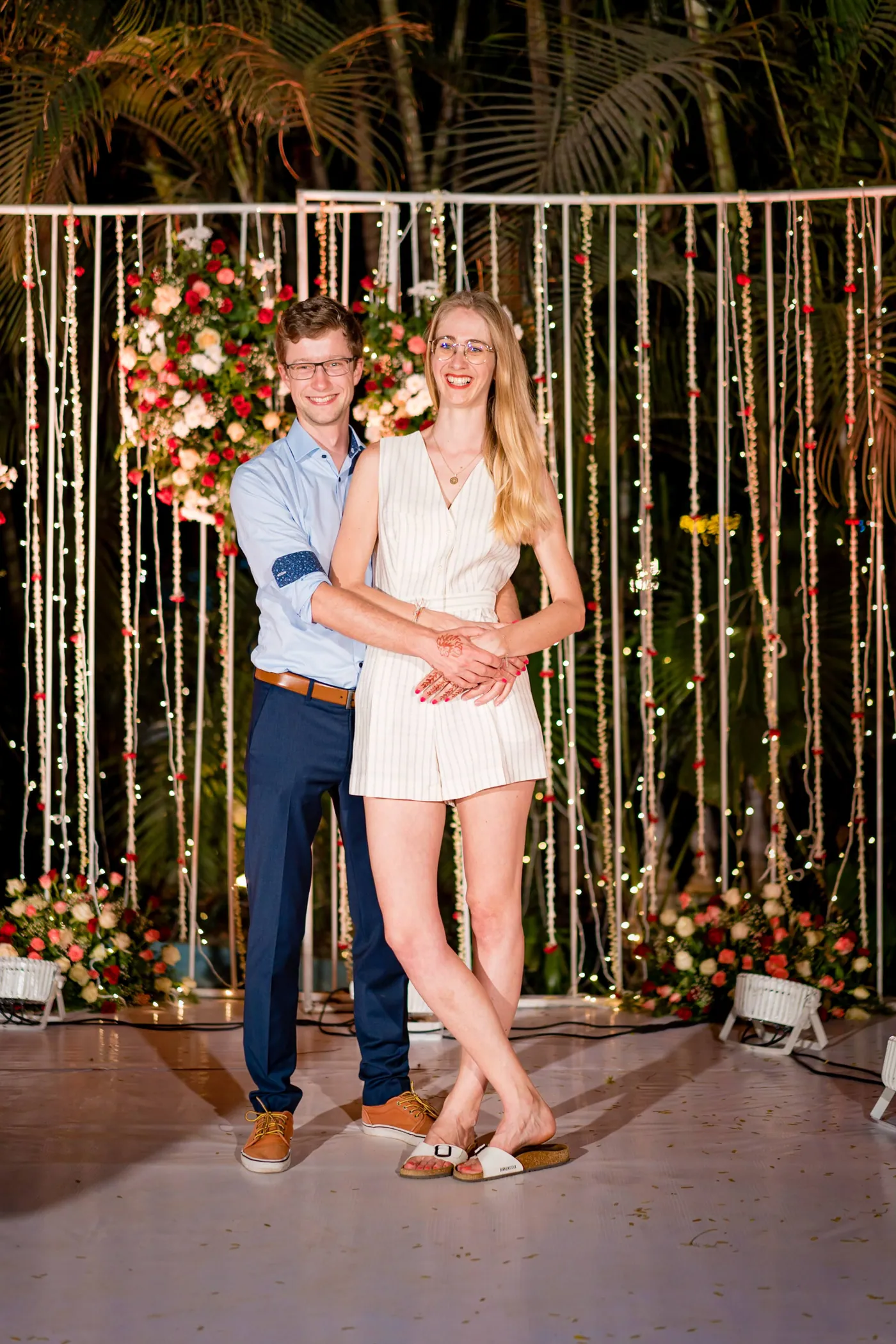 A man in a blue shirt and a woman in a white romper pose in front of the lit backdrop