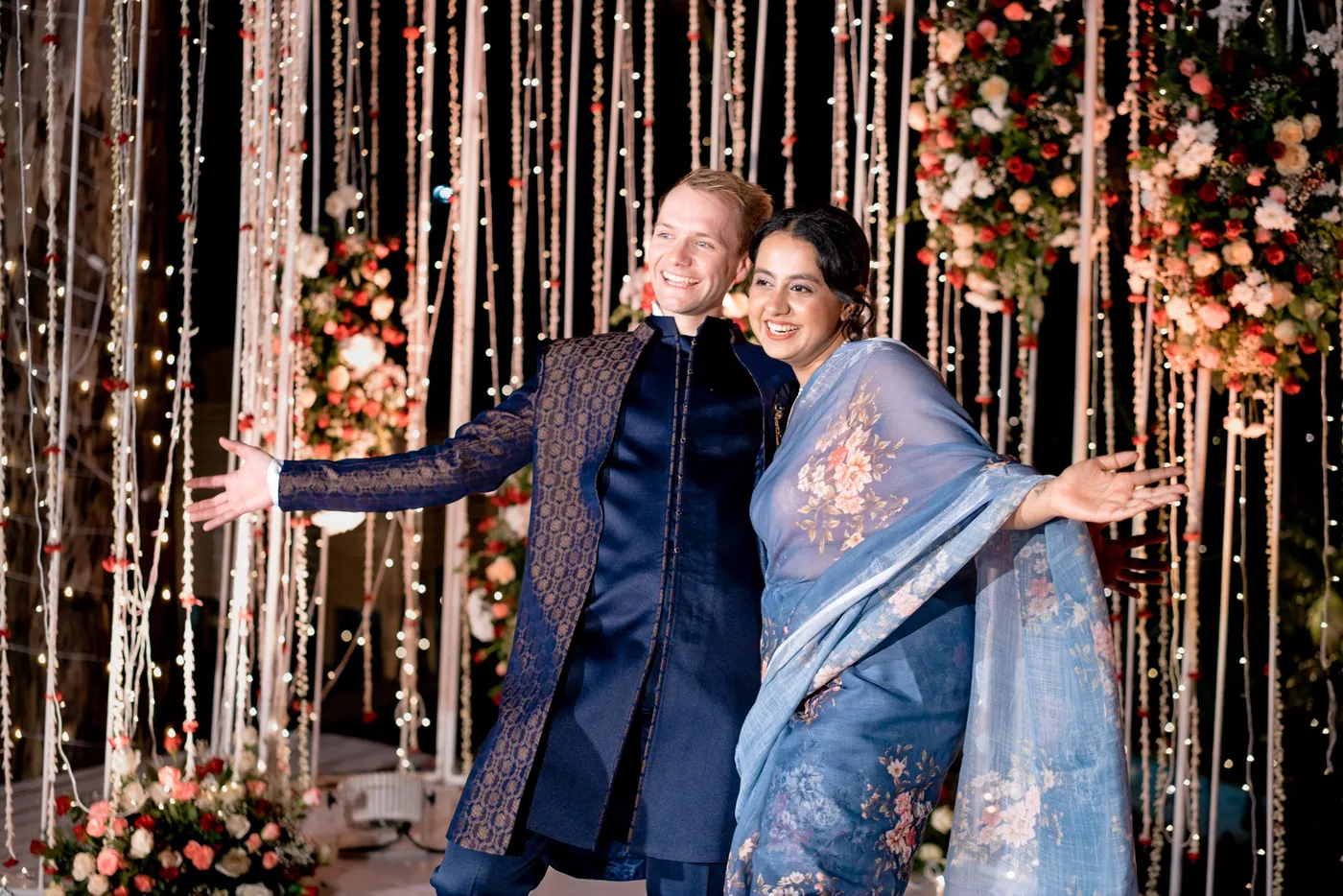 Stefan in a navy sherwani and a woman in a blue sari throw their arms wide before the lit backdrop