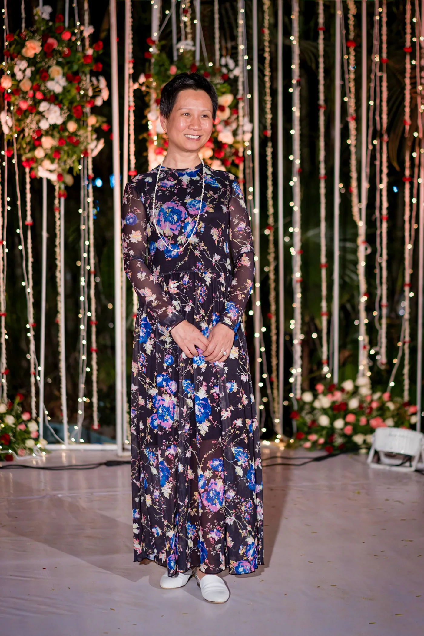 A woman in a floral jumpsuit stands for a portrait in front of the lit backdrop
