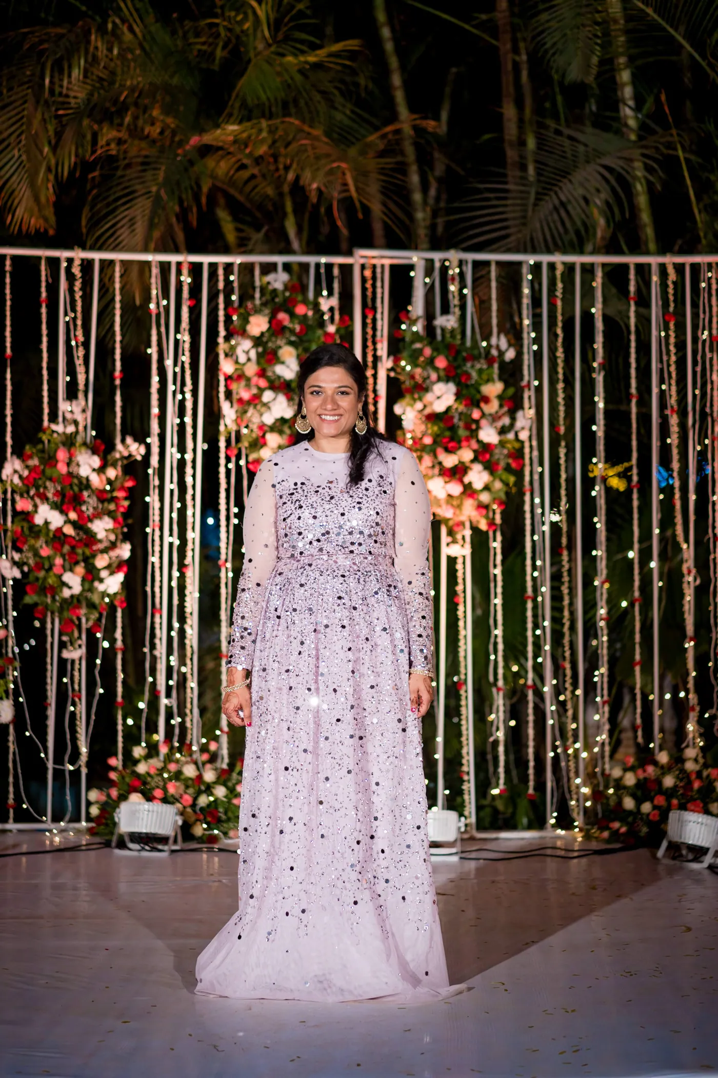 A woman in a lilac sequined gown stands alone in front of the glowing backdrop