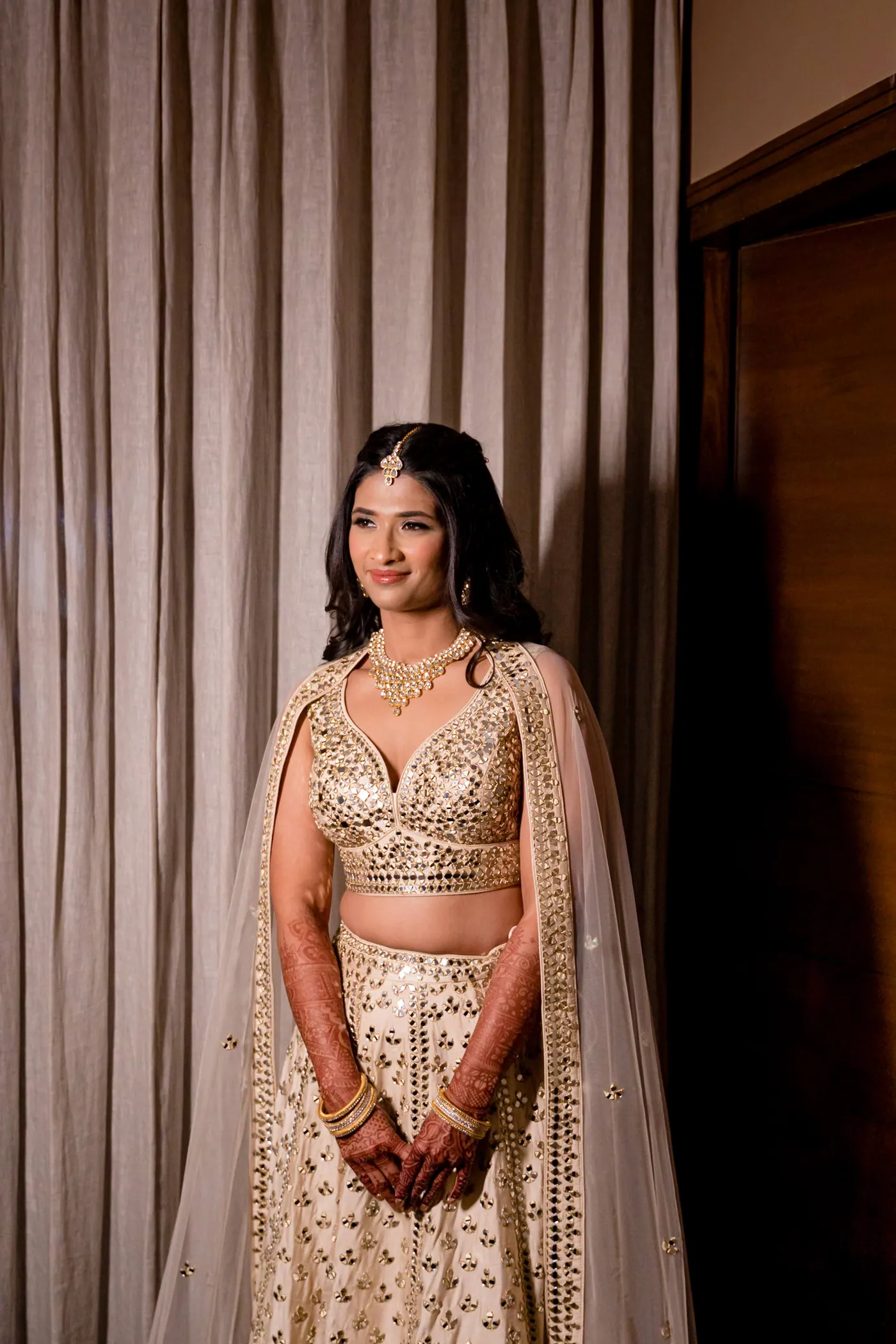 Medha stands in her cream lehenga beside heavy curtains, smiling softly past the camera