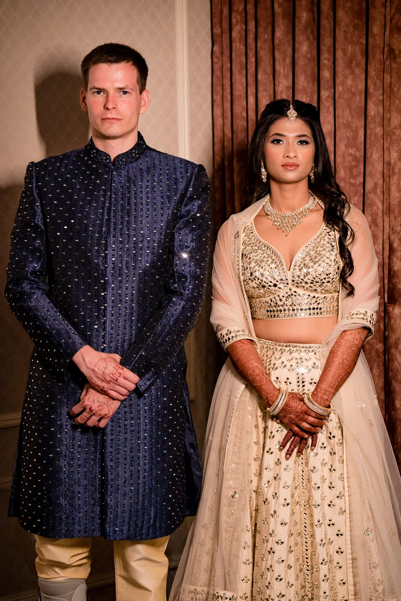 Stefan in a navy sherwani and Medha in cream stand side by side, hands clasped, in front of curtains